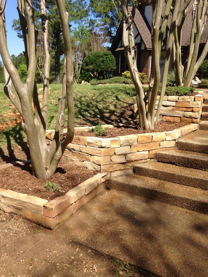 Stone retaining wall and entry steps framed by crepe myrtles and fresh planting on a sloped front yard