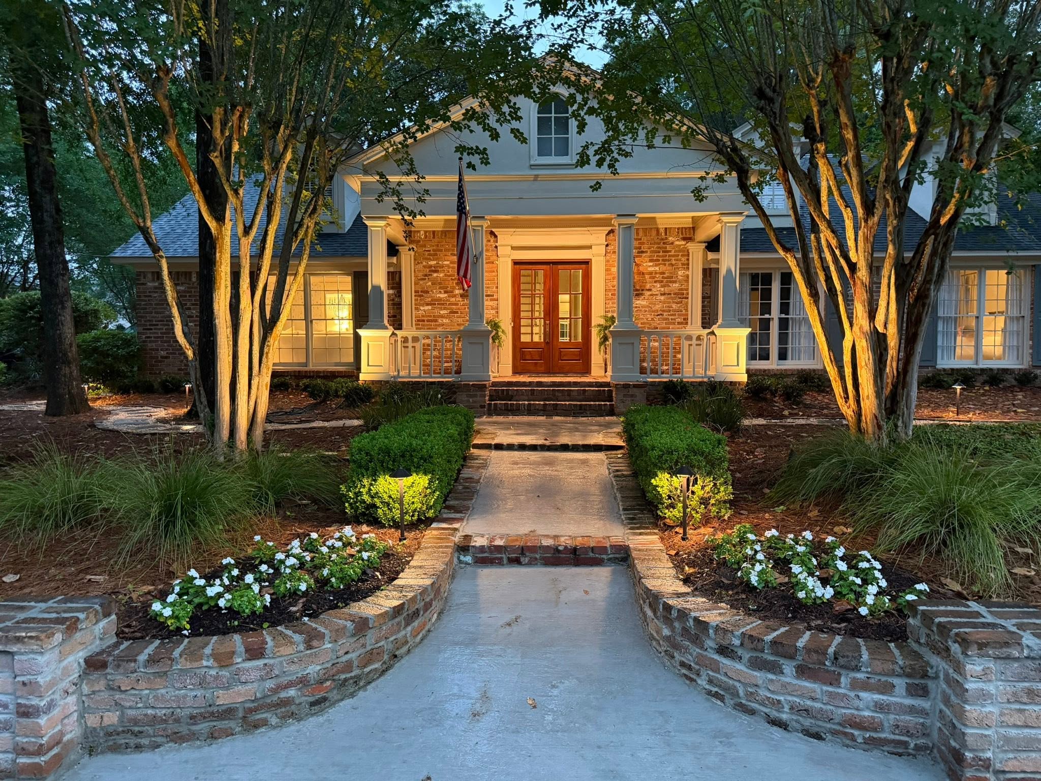 A brick home at dusk, its entry walk, flower beds and crepe myrtles lit in warm gold
