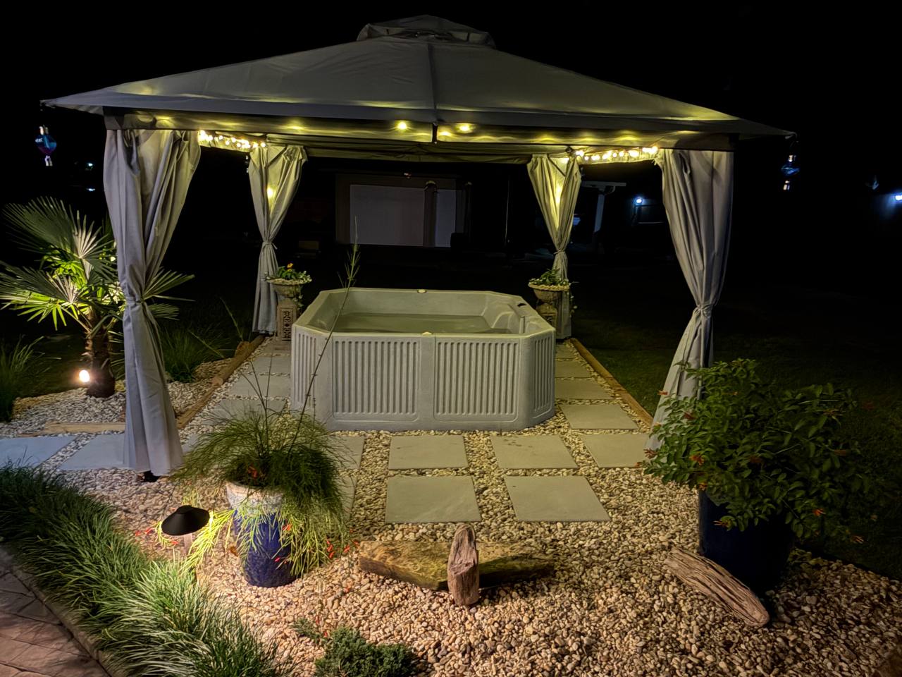 Spa tucked beneath a canopy with warm string lights, path lighting, and planted containers