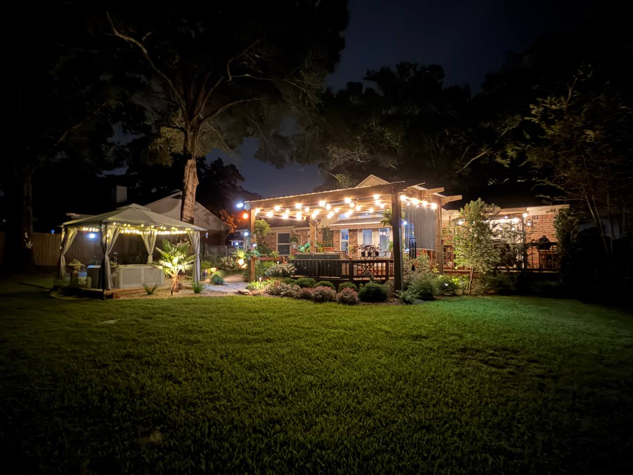 Wide evening view of a landscaped backyard with spa canopy, pergola lighting, and layered garden illumination