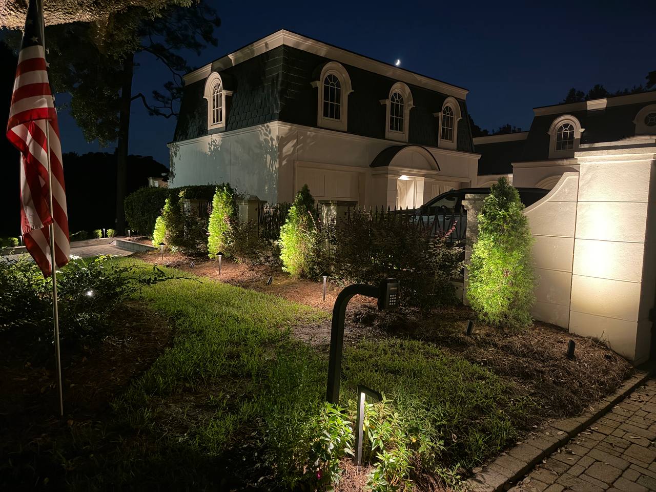 Corner of a two-story stucco home with dormer windows, an iron fence, and a row of uplit arborvitae along the drive