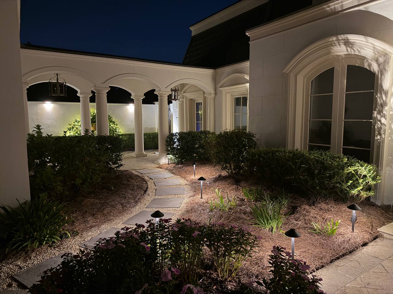Arched colonnade with hanging lanterns beside a stepping-stone path lit by mushroom path lights through a planting bed