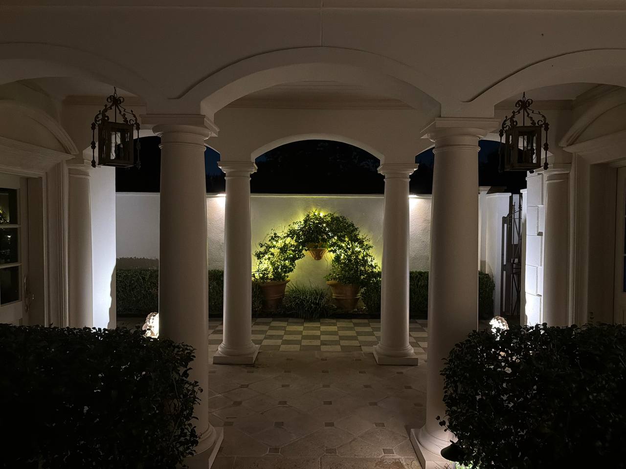 View through a colonnade of columns and hanging lanterns to a checkerboard-tile courtyard with a backlit circular vine arch on the far wall