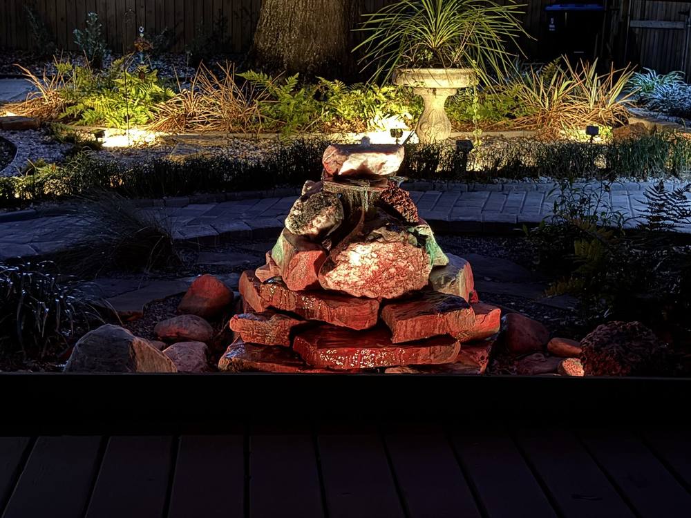 Stacked stone water feature with a red glazed urn and trailing greenery beside a riverfront lawn