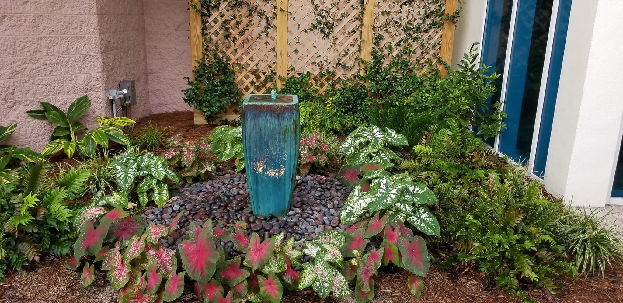 Blue glazed urn fountain bubbling over black river stone with caladiums, ferns, lattice, and entry windows behind it